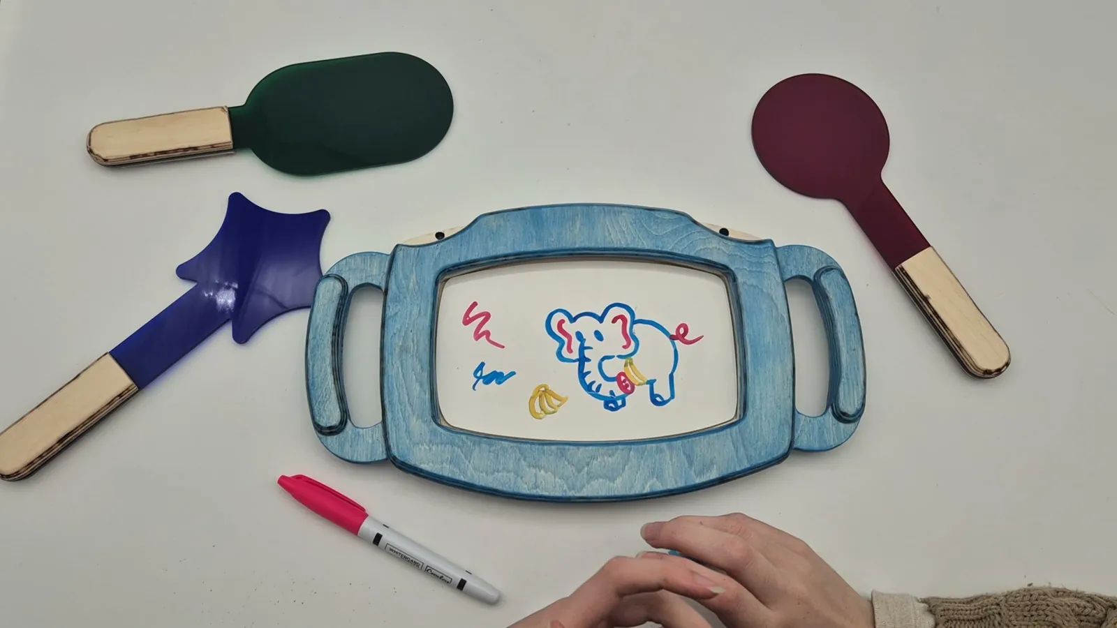 Still from the product video: the painted frame with an elephant drawing, the three paddles and markers